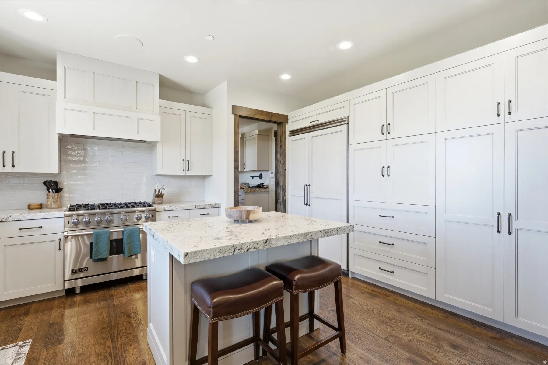 Gourmet kitchen featuring a built-in stainless steel range, white shaker cabinetry, an integrated panel-ready refrigerator, wood-finish flooring, and a central island with a stone countertop