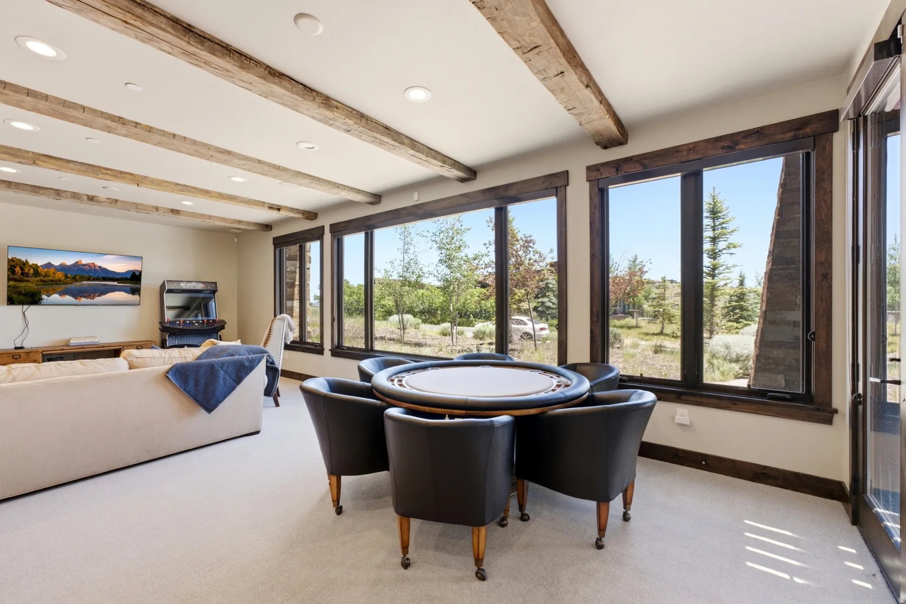 Spacious interior featuring exposed wood ceiling beams, recessed lighting, a wall of large windows with dark wood trim, and light-colored carpeting