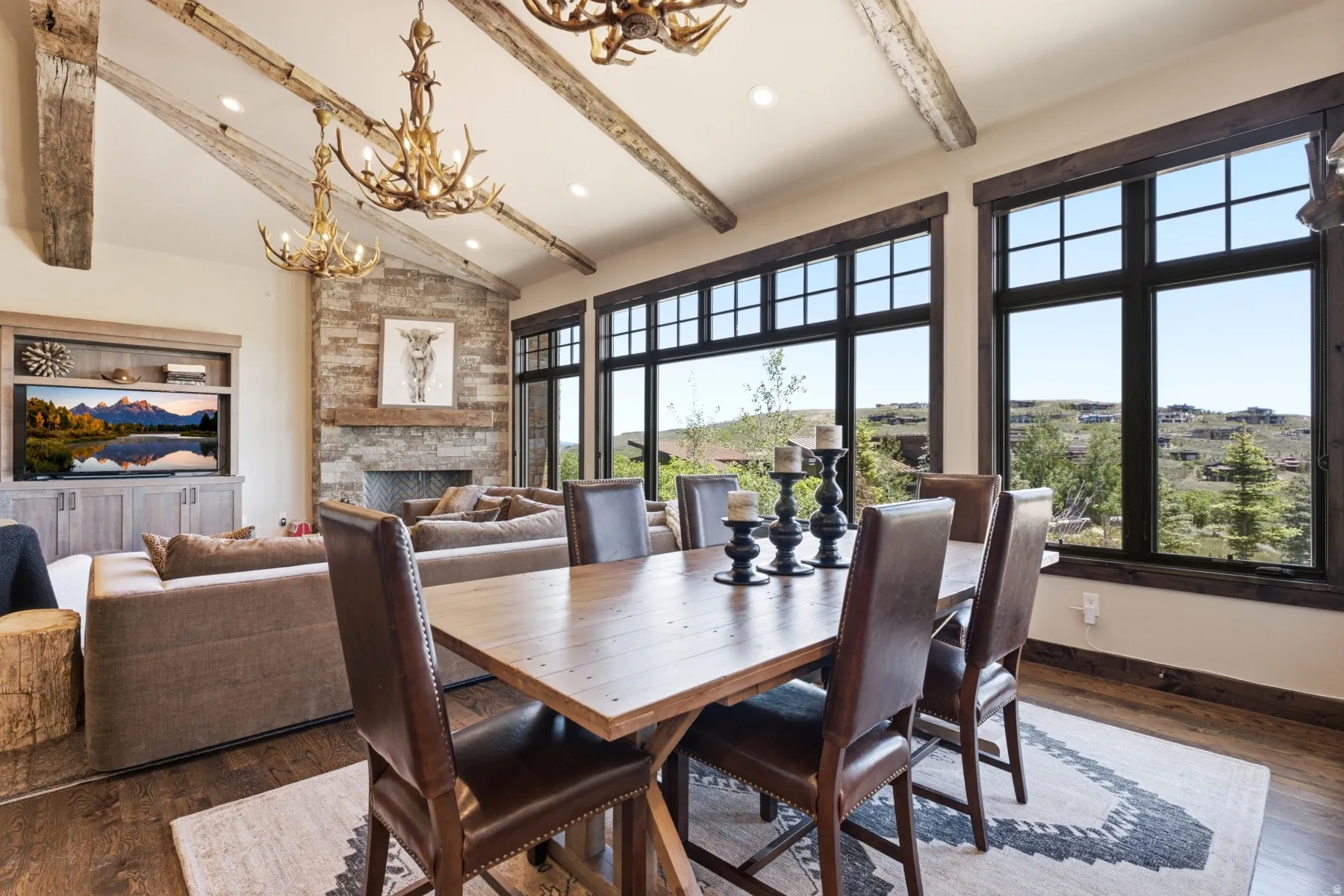 Open-concept living and dining area featuring a vaulted ceiling with exposed wood beams, a stone-faced fireplace with a wood mantel, a built-in media center, and expansive windows with dark frames