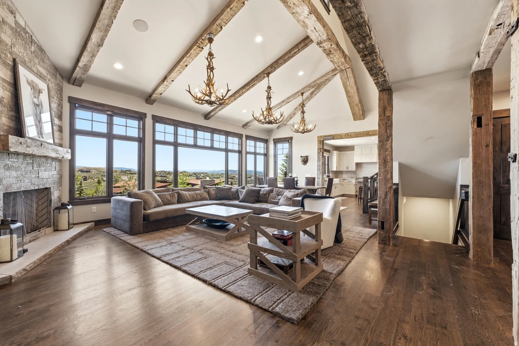 Vaulted ceilings with exposed wood beams define this expansive room featuring large picture windows, wood-finish flooring, a stone fireplace, and built-in antler-style chandeliers