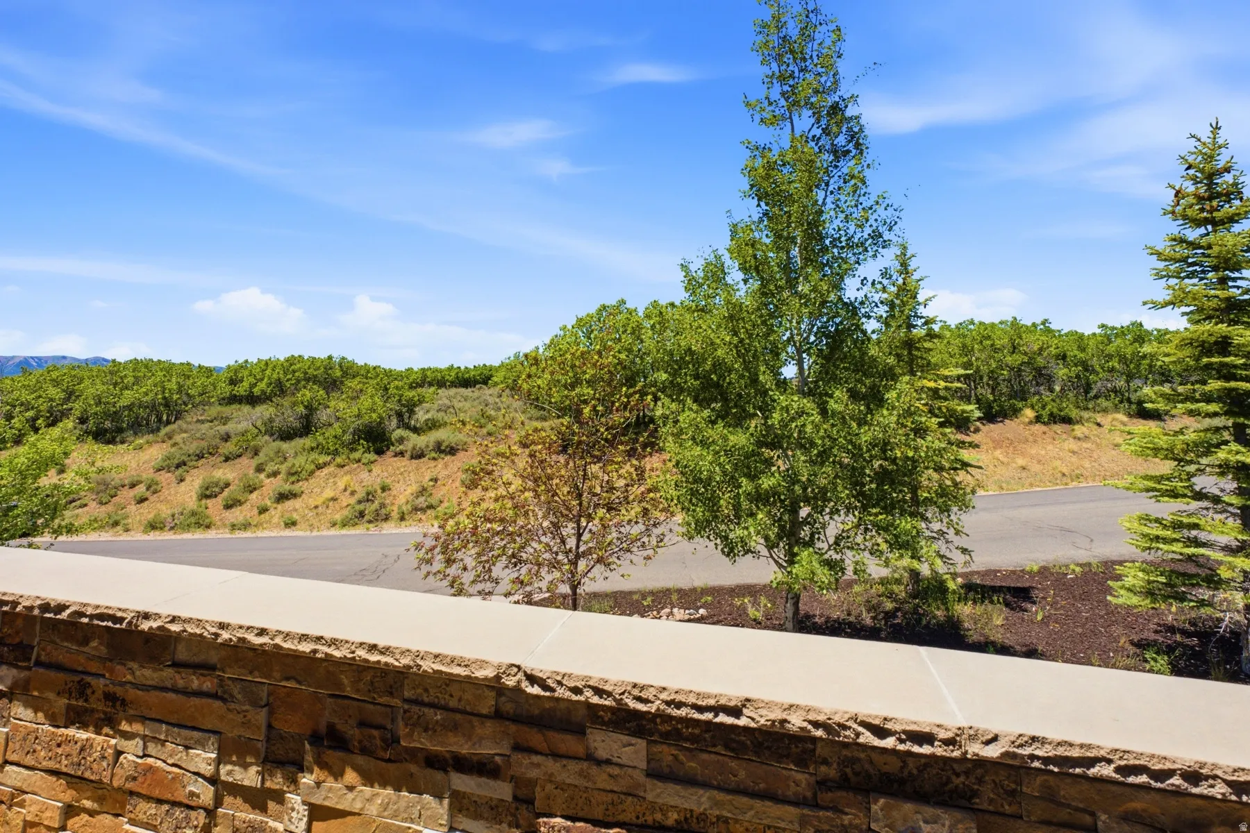 Stacked stone wall with a smooth coping, showcasing a natural landscape with varied foliage and a clear blue sky