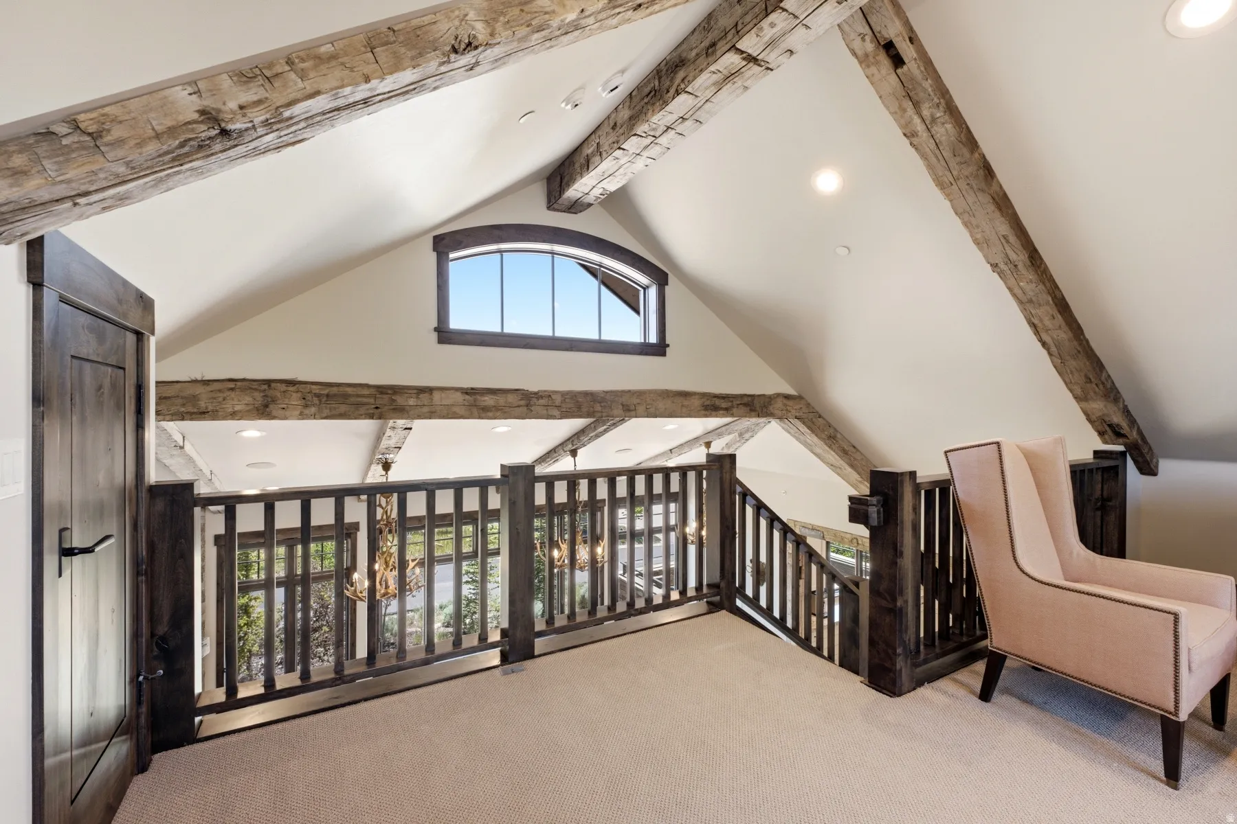 Upper-level landing featuring a vaulted ceiling with exposed wood beams, a custom arched window, and dark wood railings overlooking the level below