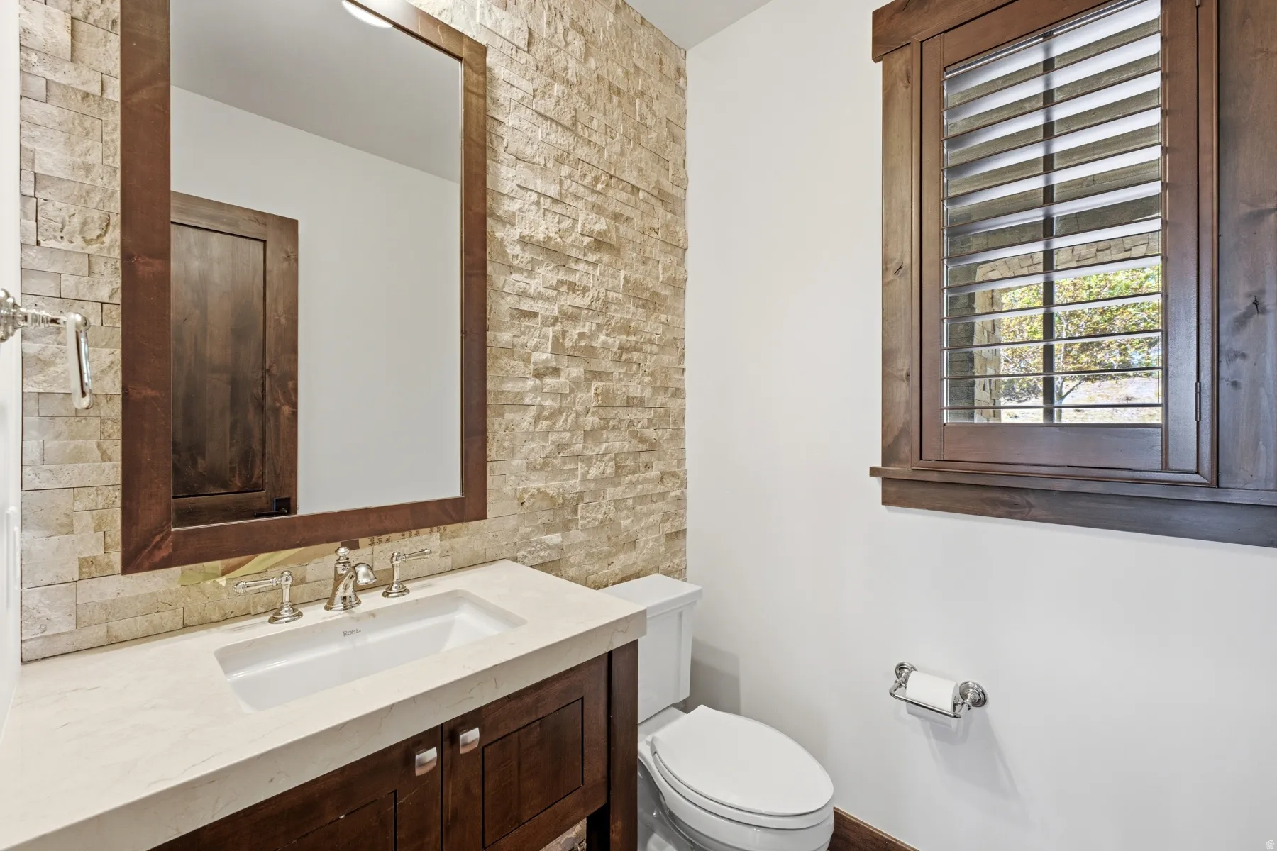 Bathroom featuring a stone accent wall, dark wood vanity with a light-toned countertop, and an undermount sink with a polished chrome faucet