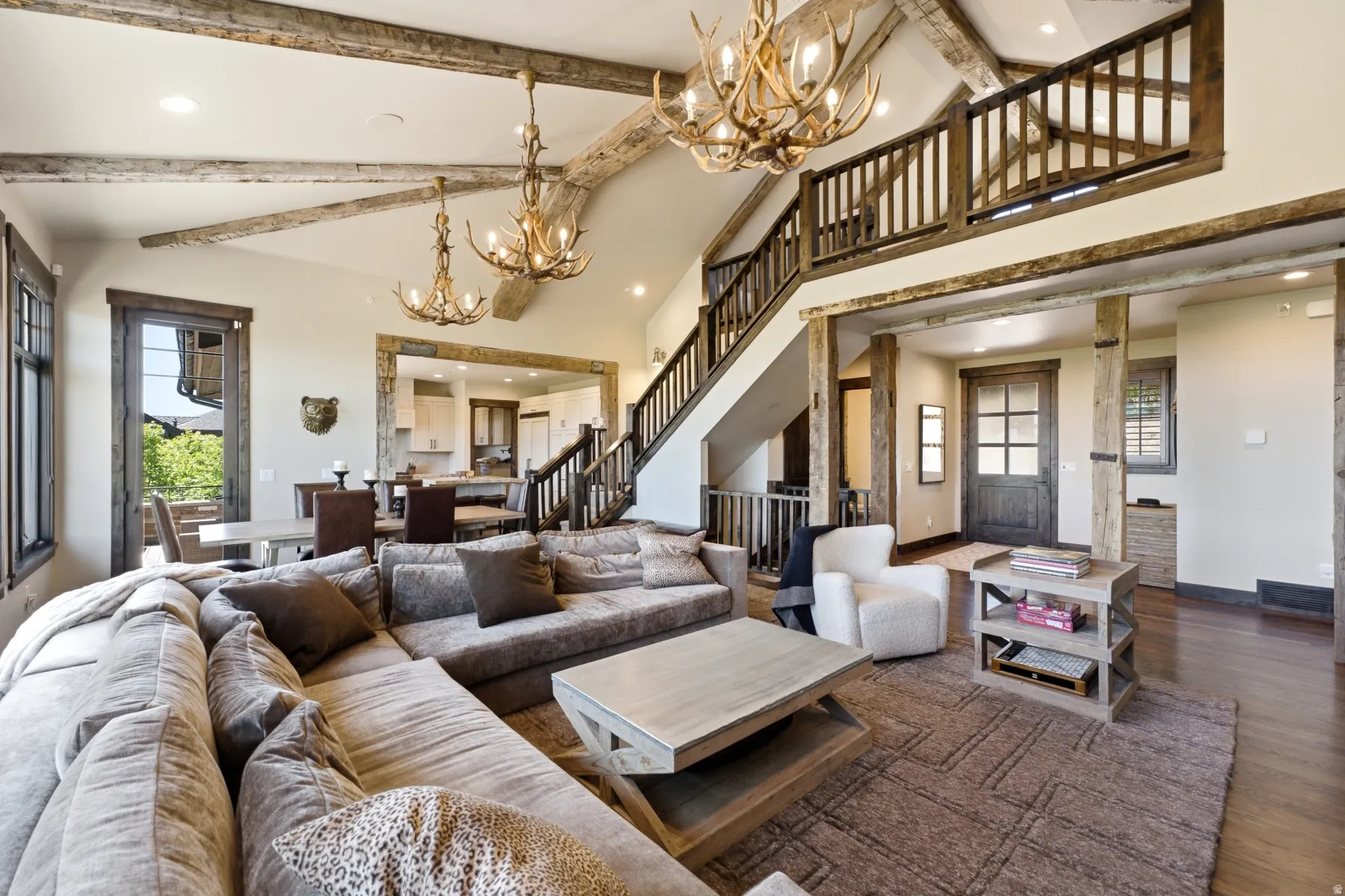 Vaulted ceilings with exposed wood beams, antler chandeliers, a multi-level staircase with wood railing, and dark wood trim throughout