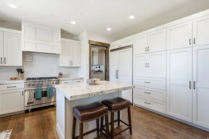 Gourmet kitchen featuring a built-in stainless steel range, white shaker cabinetry, an integrated panel-ready refrigerator, wood-finish flooring, and a central island with a stone countertop