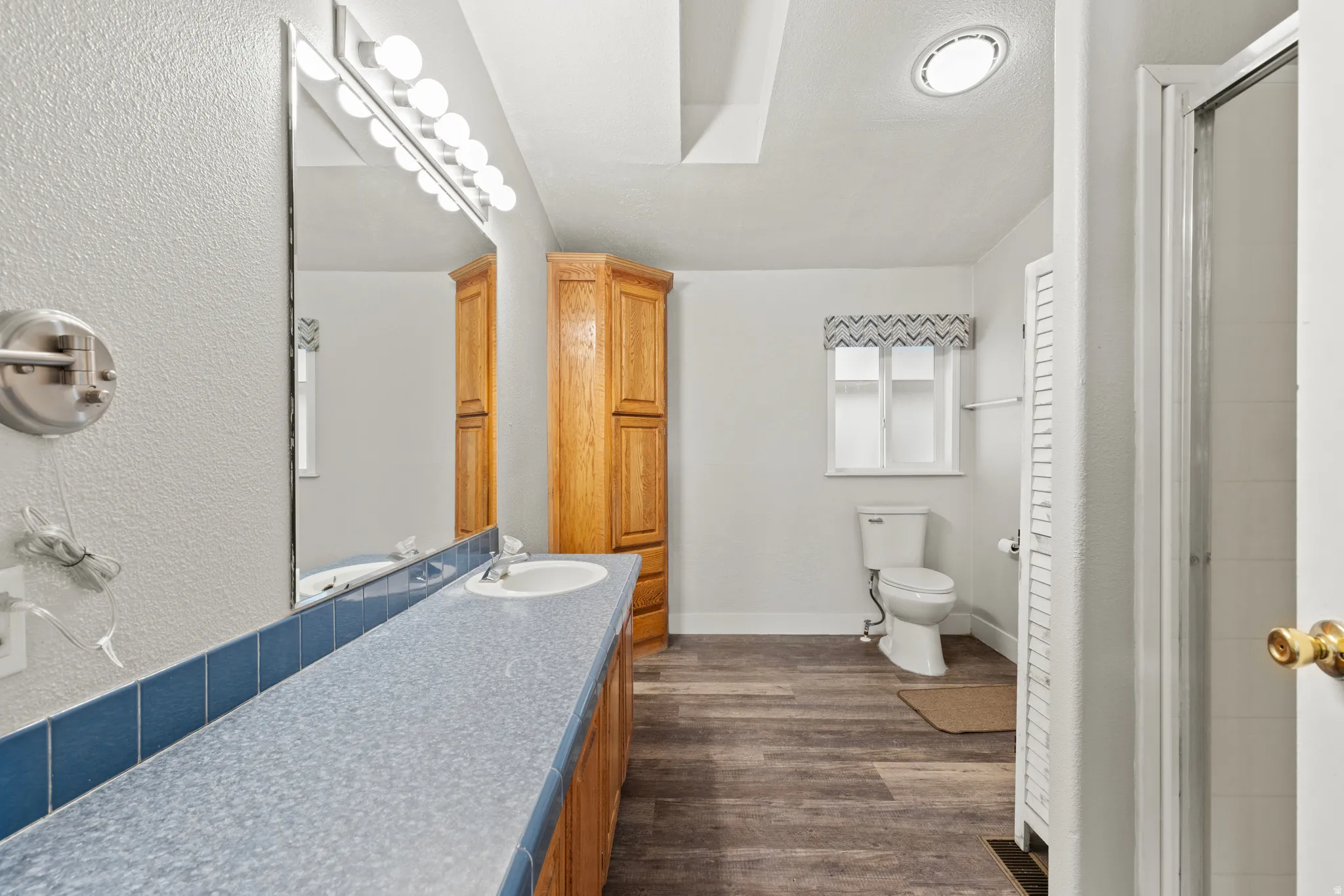 Spacious bathroom featuring a long vanity with a light blue countertop and integrated sink, wood-finish flooring, a corner built-in wood cabinet, and a shower enclosure with a glass door