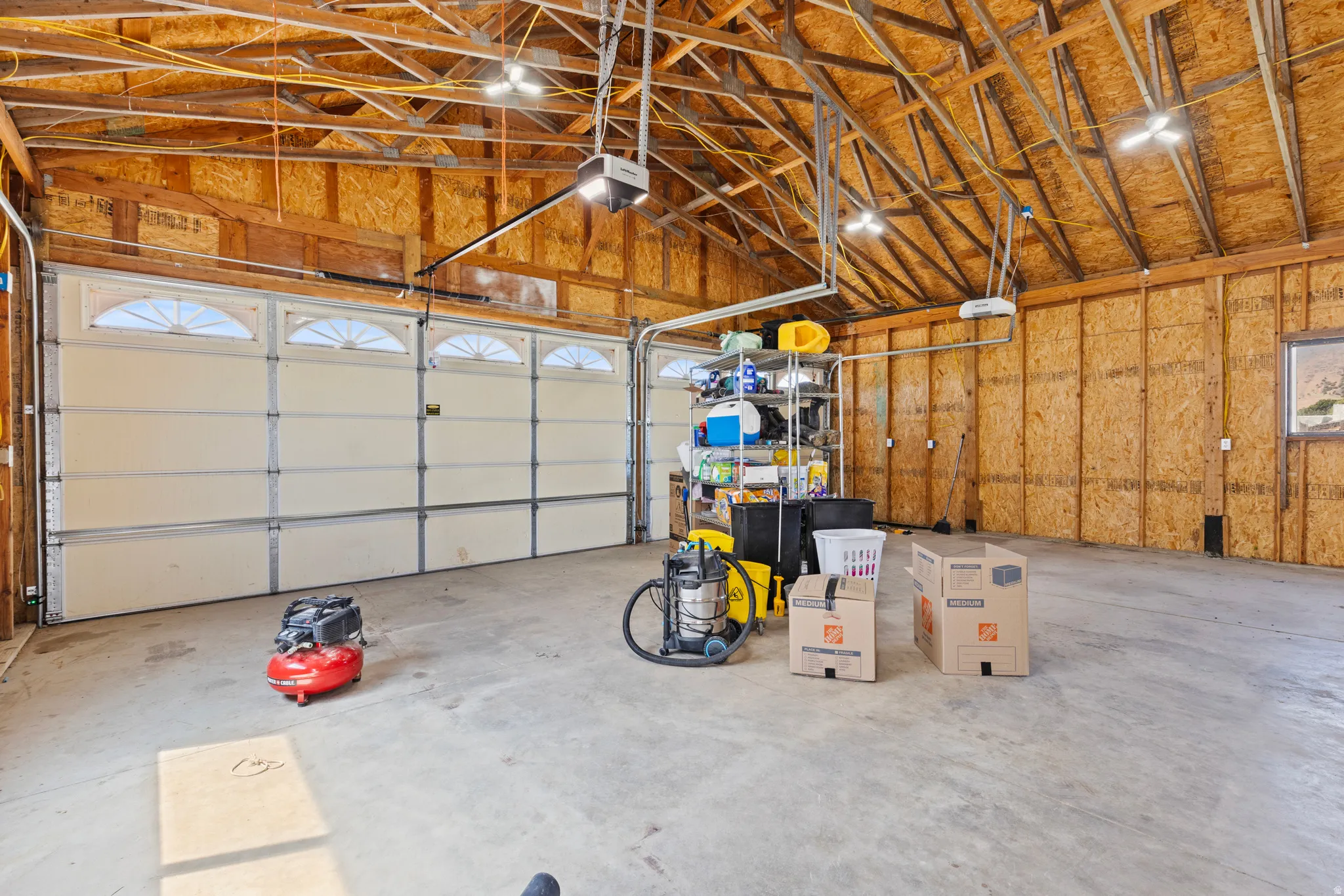 Spacious garage featuring exposed wood framing, multiple garage doors with arched window panels, concrete flooring, and overhead lighting fixtures