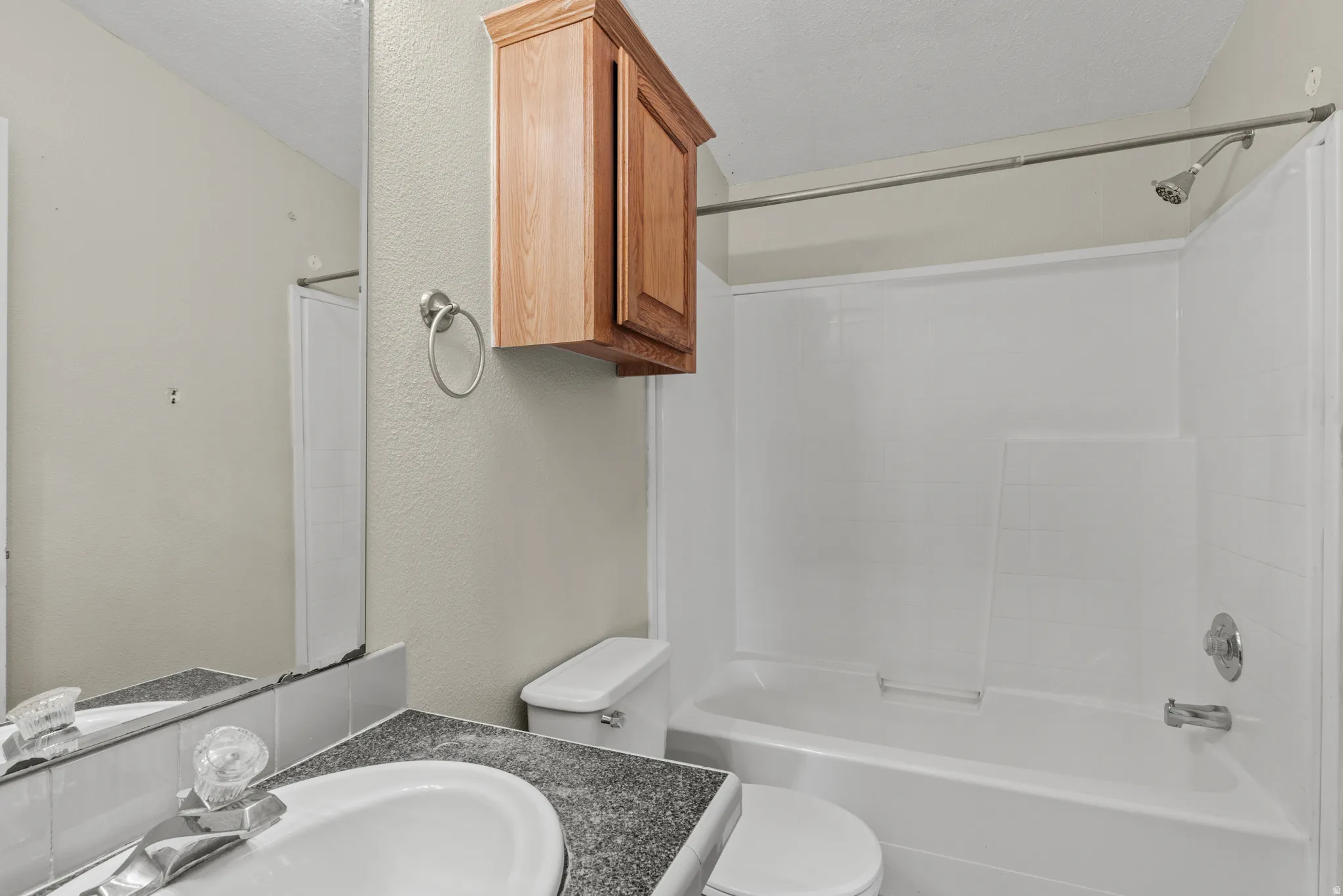 Bathroom featuring a white bathtub and shower surround, a chrome showerhead, a toilet, a light wood-finish storage cabinet, and a vanity with a white sink and dark speckled countertop