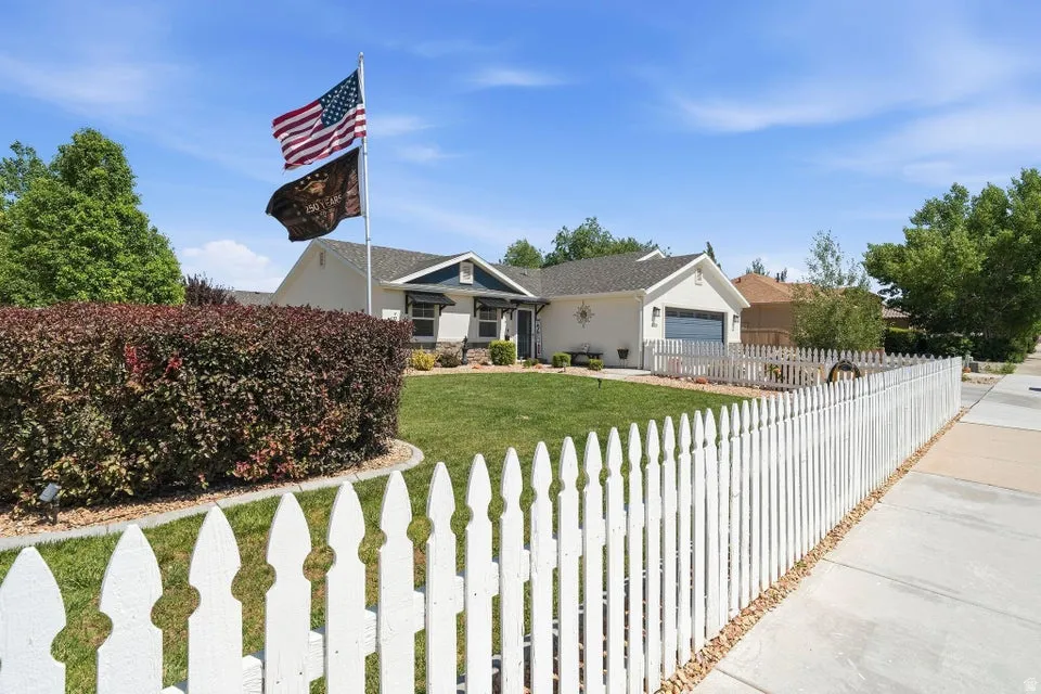 Front exterior featuring a white picket fence, a manicured lawn, established shrubbery, an attached garage, and a concrete sidewalk