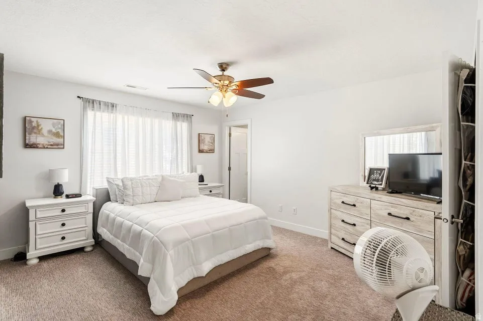 Carpeted bedroom with a ceiling fan, white walls, and a large window with blinds and sheer curtains