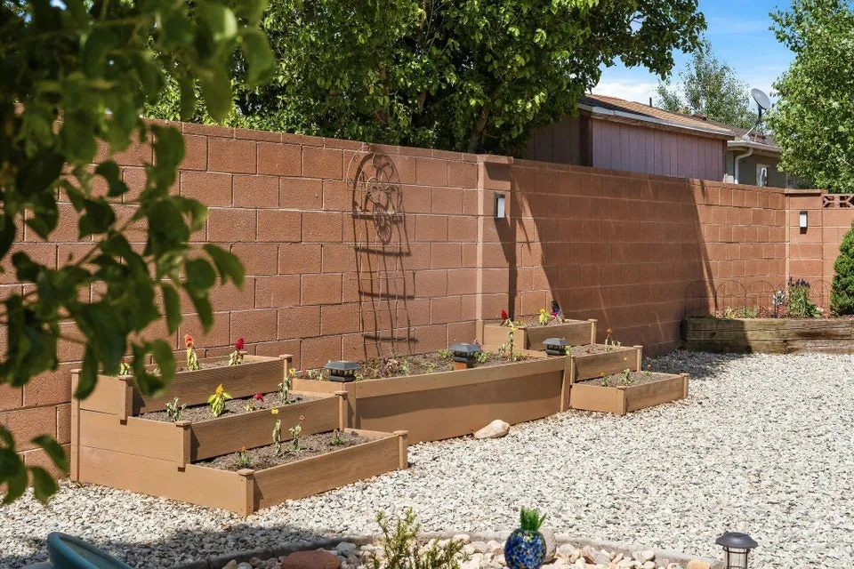Gravel-surfaced backyard featuring tiered planters with young plantings and a sturdy block wall with integrated lighting