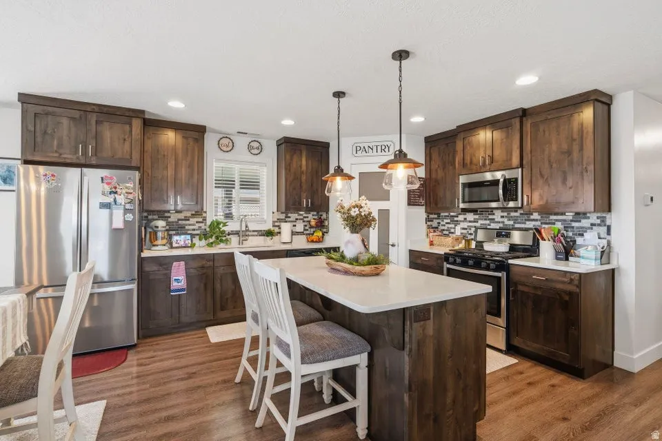 Kitchen featuring wood-finish flooring, dark wood cabinetry, stainless steel appliances, a mosaic tile backsplash, and a central island with a light-colored countertop