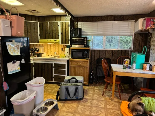 Kitchen featuring wood-paneled walls, a double basin sink, ample upper and lower cabinetry, a built-in range hood, and a window with horizontal blinds
