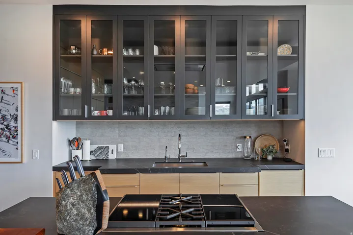 Sleek kitchen featuring black frame glass-front upper cabinetry with integrated lighting, wood-finish lower cabinets, a black countertop with an integrated sink, a gray textured backsplash, and a black cooktop