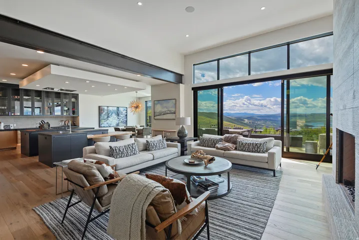 Expansive open-concept living area featuring a wood-finish flooring, a contemporary fireplace with a stone surround, and floor-to-ceiling sliding glass doors offering panoramic mountain and lake scenery