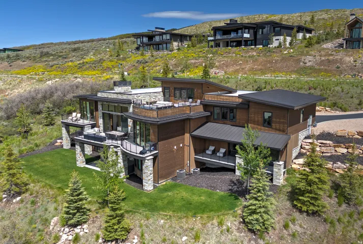 Modern multi-level residence featuring extensive stone and wood siding, multiple exterior balconies with glass railings, a flat roofline, and integrated landscaping with mature trees