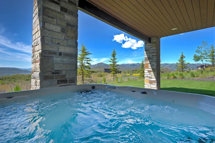 Outdoor spa situated under a covered patio with stone columns, featuring a clear sky, distant mountains, and a grassy area