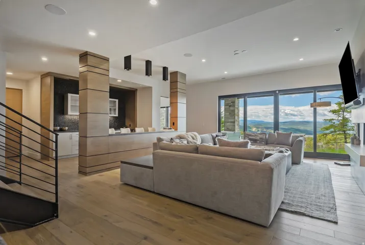 Open-concept living area with wood-finish flooring, a contemporary wet bar featuring dark tile backsplash, and expansive floor-to-ceiling sliding glass doors revealing mountain scenery