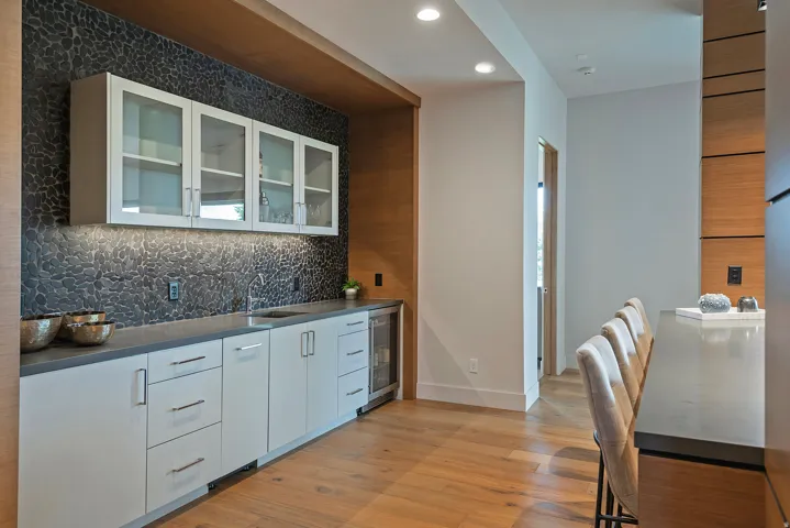 Built-in wet bar featuring a textured dark stone backsplash, under-cabinet lighting, and a stainless steel beverage refrigerator