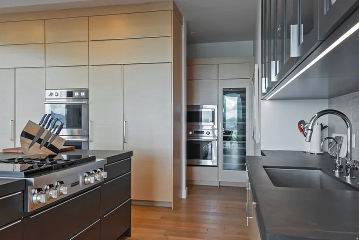 Modern kitchen featuring a large island with a gas range, integrated stainless steel appliances, light wood-finish cabinetry, dark countertops, and wood-finish flooring