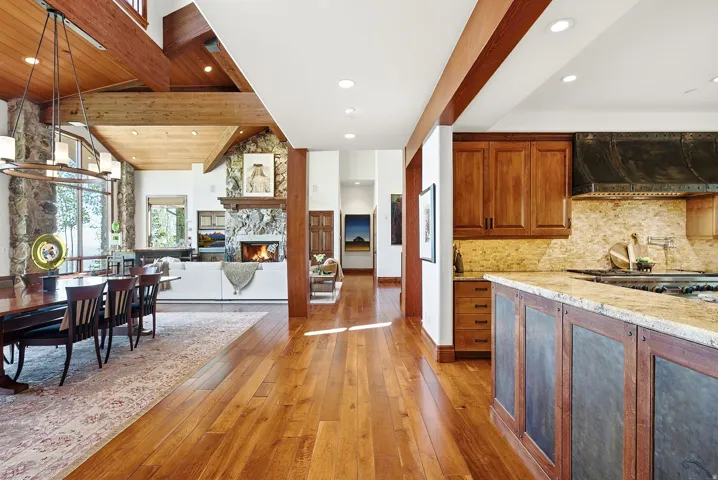 Open-concept living space featuring wood-finish flooring, exposed wood beams, a stone-clad fireplace with a wood mantle, and large windows