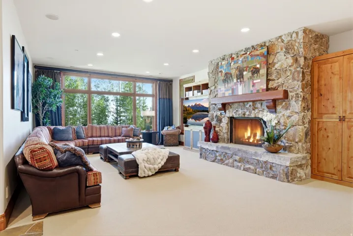 Spacious living area featuring a stone-clad fireplace with a substantial timber mantel, large picture windows, recessed ceiling lighting, and built-in wood cabinetry