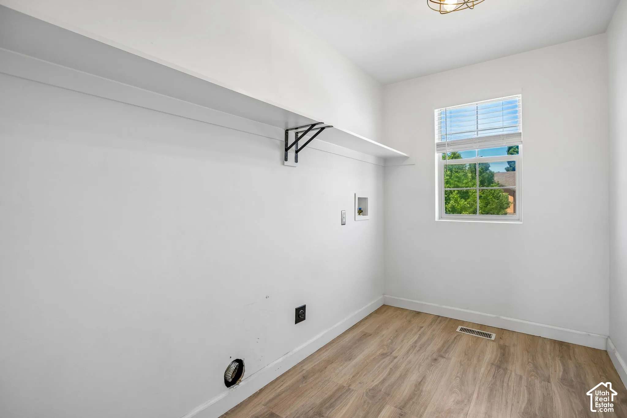 Laundry area featuring a window with blinds, a built-in shelf with black brackets, wood-finish flooring, and white baseboards