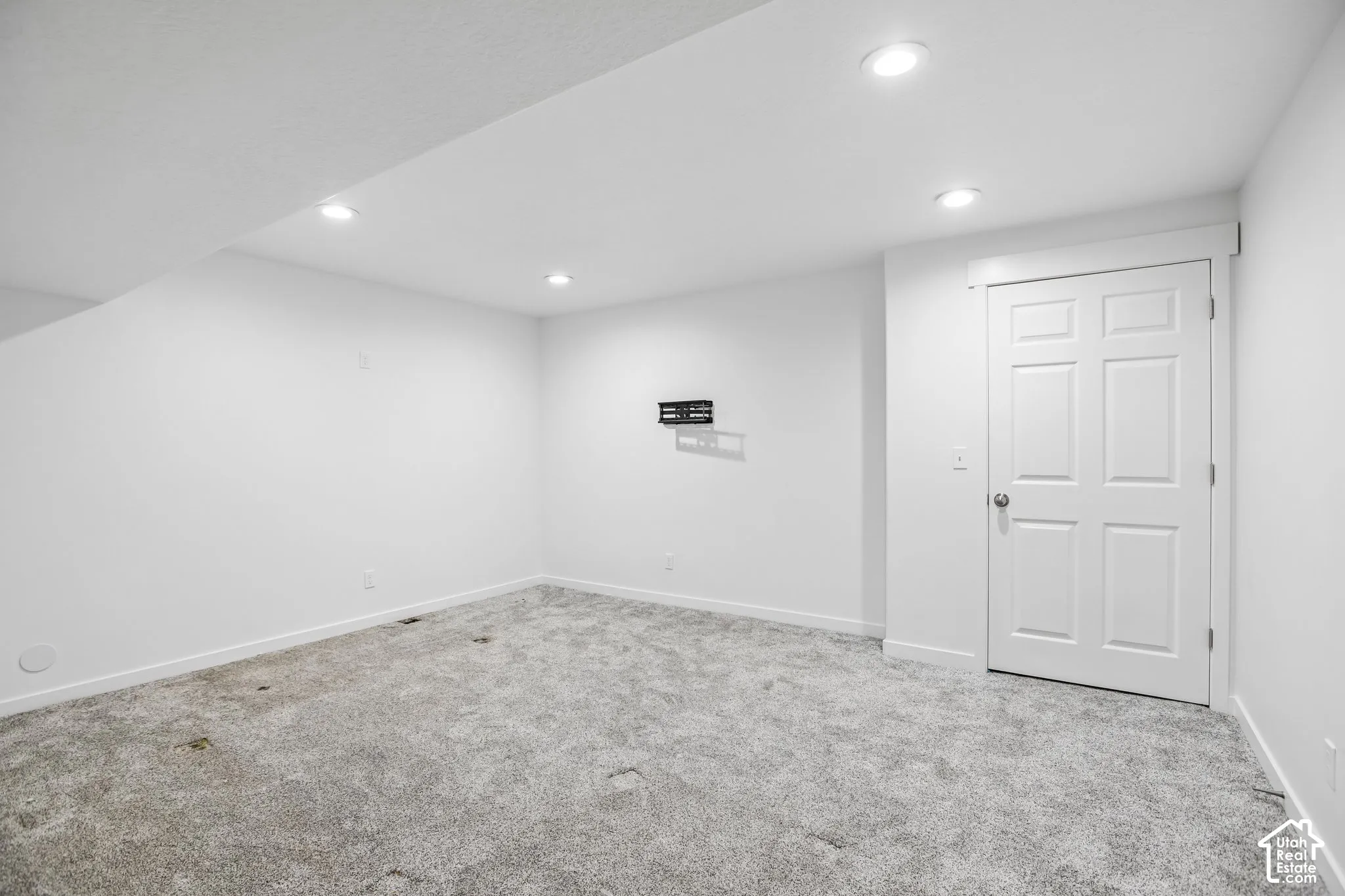 Carpeted room featuring recessed lighting, a white six-panel door with silver hardware, white baseboards, and a wall-mounted bracket