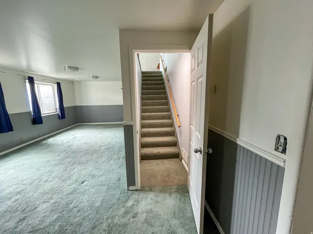 Carpeted staircase with wooden handrail opening to a spacious room featuring a light-colored carpet, multiple windows, and two-tone wall treatment with lower paneling