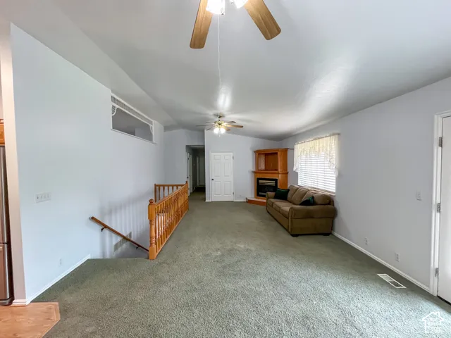 Spacious carpeted living area featuring a corner fireplace with wood surround, ceiling fans, and a window with blinds