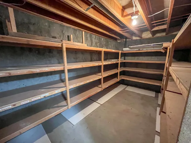 Basement storage area featuring extensive built-in wooden shelving, exposed ceiling joists, and concrete walls