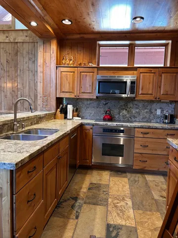 Kitchen featuring wood cabinetry, stone countertops, a double basin sink with a gooseneck faucet, stainless steel appliances, and multi-toned stone tile flooring