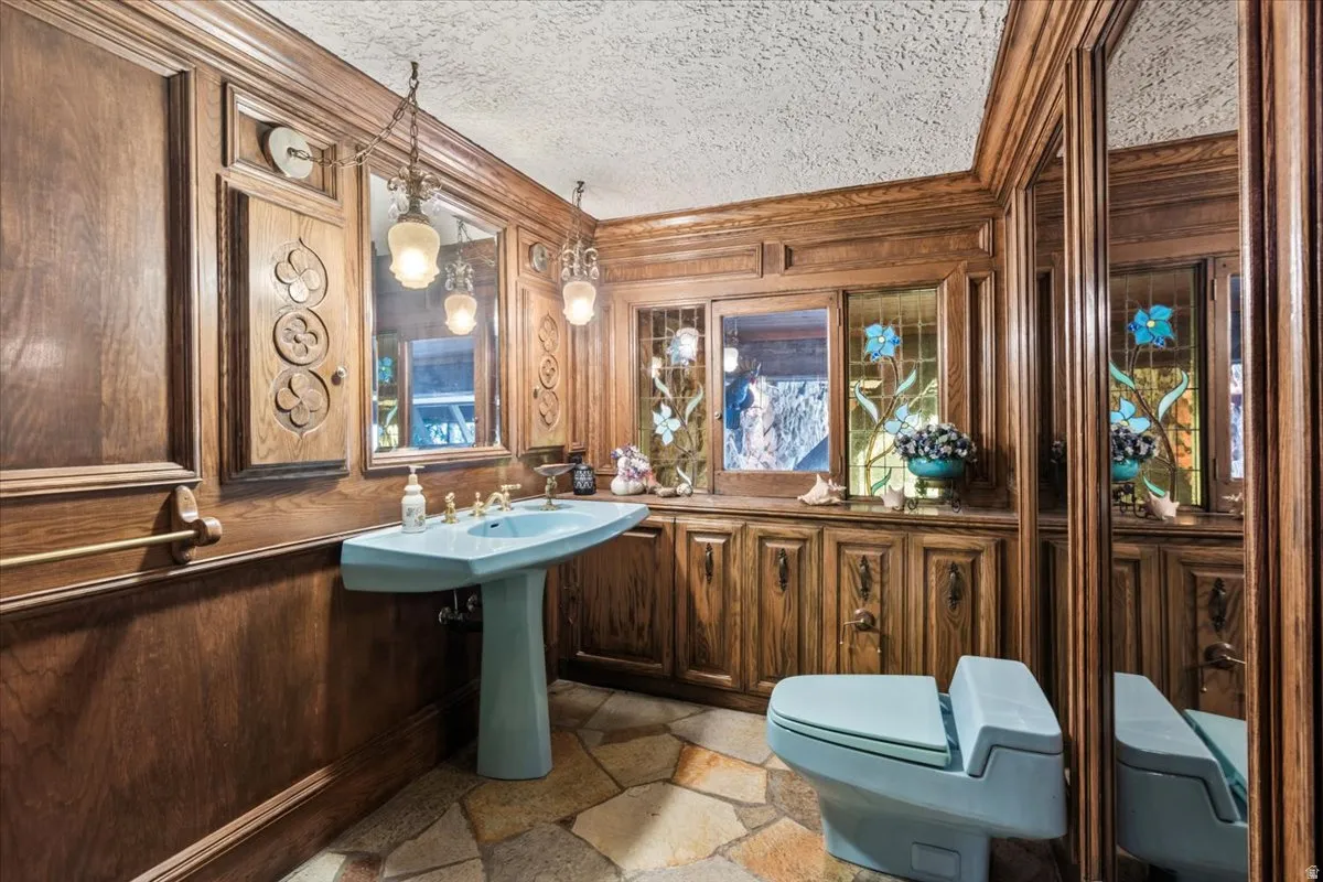 Bathroom featuring extensive wood paneling, a light blue pedestal sink with brass fixtures, a matching light blue toilet, and stained glass window accents