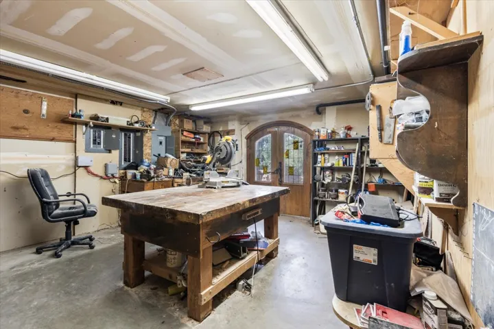 Spacious workshop area featuring concrete flooring, exposed ceiling beams, built-in shelving, ample fluorescent lighting, and double arched wood doors with glass inserts