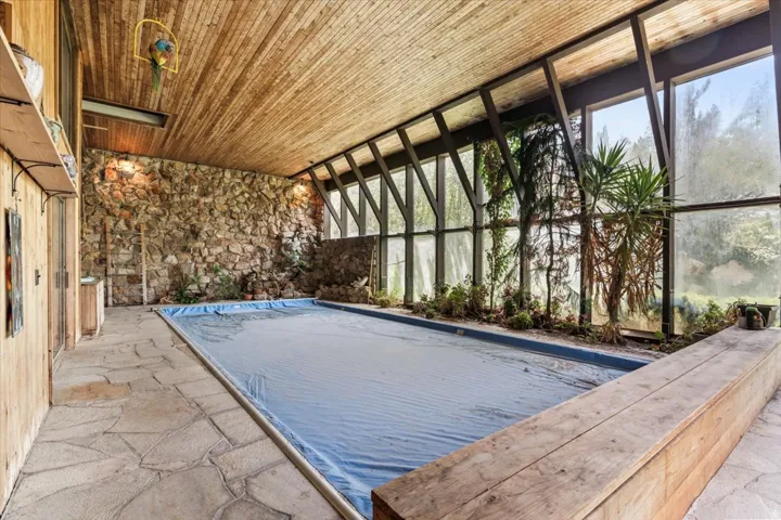 Indoor pool featuring a slatted wood ceiling, natural stone accent wall, flagstone flooring, and a full-height window wall with integrated planters