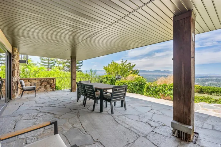 Expansive flagstone patio featuring a covered outdoor area, stone wall with metal railing, and a distant mountain landscape