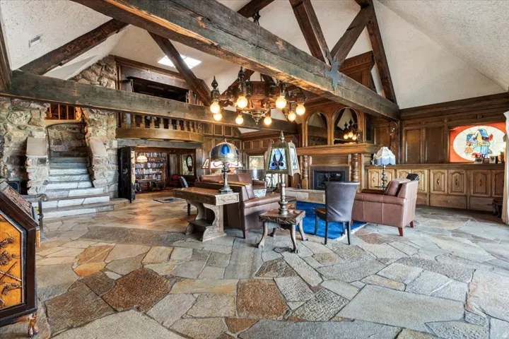 Vaulted ceilings with exposed wood beams, flagstone flooring, a stone staircase with a wrought-iron railing, a stone fireplace with a wood mantel, and wood paneling with integrated cabinetry