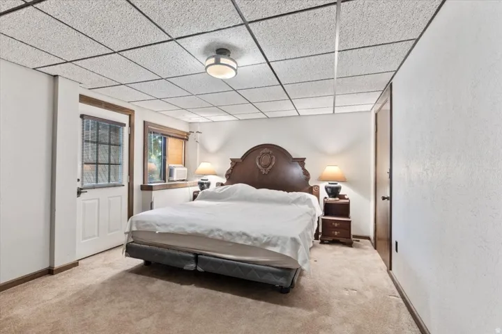 Carpeted room featuring a white paneled door with window, a window with exterior visibility, a drop ceiling, and a built-in wall air conditioning unit