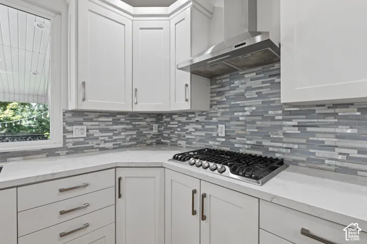 White shaker cabinetry, light-toned countertops, a gas range with stainless steel hood, and a linear mosaic tile backsplash