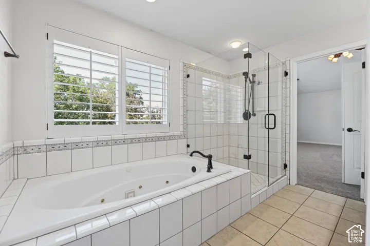 Built-in jetted tub surrounded by white tile, glass-enclosed shower with white tile, plantation shutters, and light-toned floor tiles