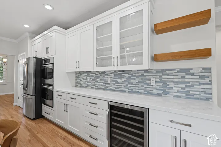 White cabinetry with glass-front uppers, stainless steel appliances, a mosaic tile backsplash, built-in wine refrigeration, and light wood-finish flooring