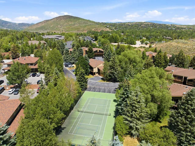 Full-size tennis court featuring a hard surface with green and blue playing areas, surrounded by mature trees