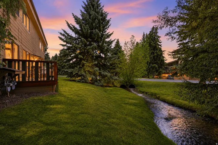 Rear exterior featuring a wood deck, verdant lawn, flowing stream, and mature evergreen trees
