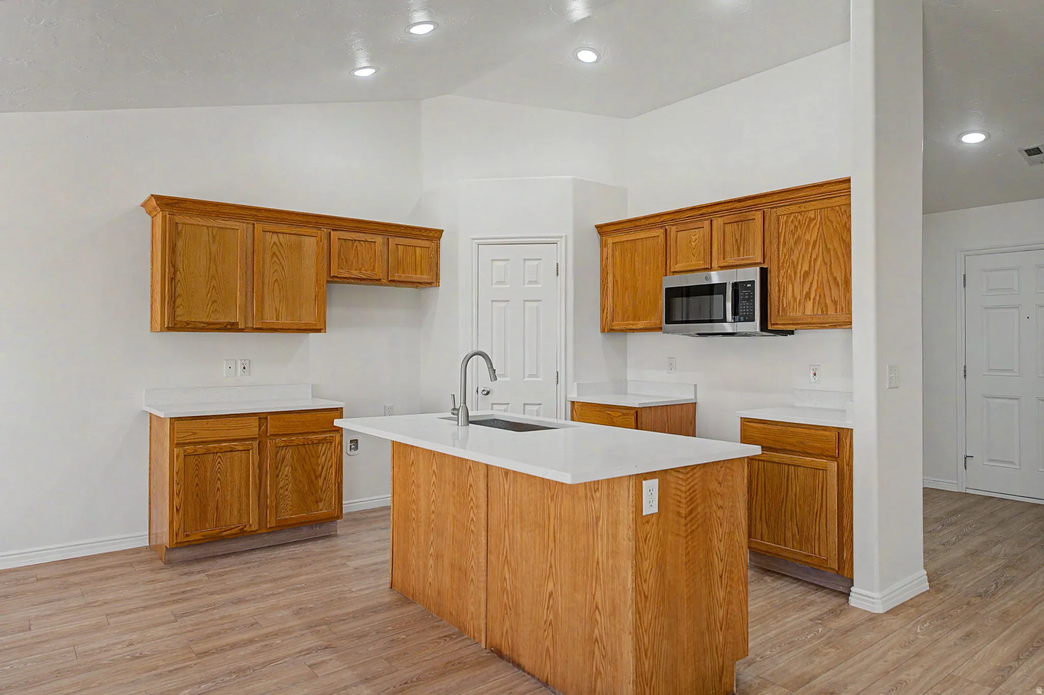Kitchen featuring an island with a sink, stainless steel microwave, oak-finish cabinetry, light-toned countertops, and recessed lighting