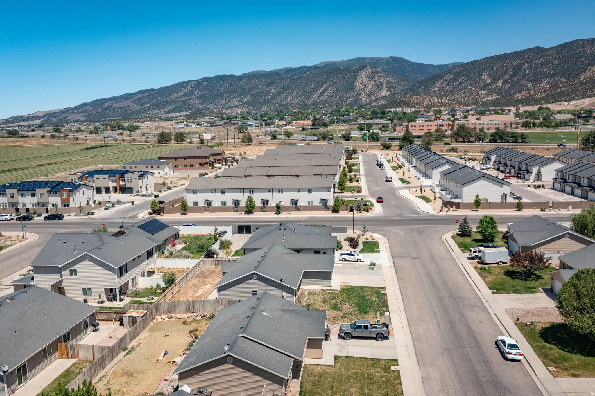 Aerial perspective of a developed residential area featuring paved streets, individual homes with gray roofs and light-colored siding, and rows of multi-unit townhomes with dark roofs and light-colored exteriors