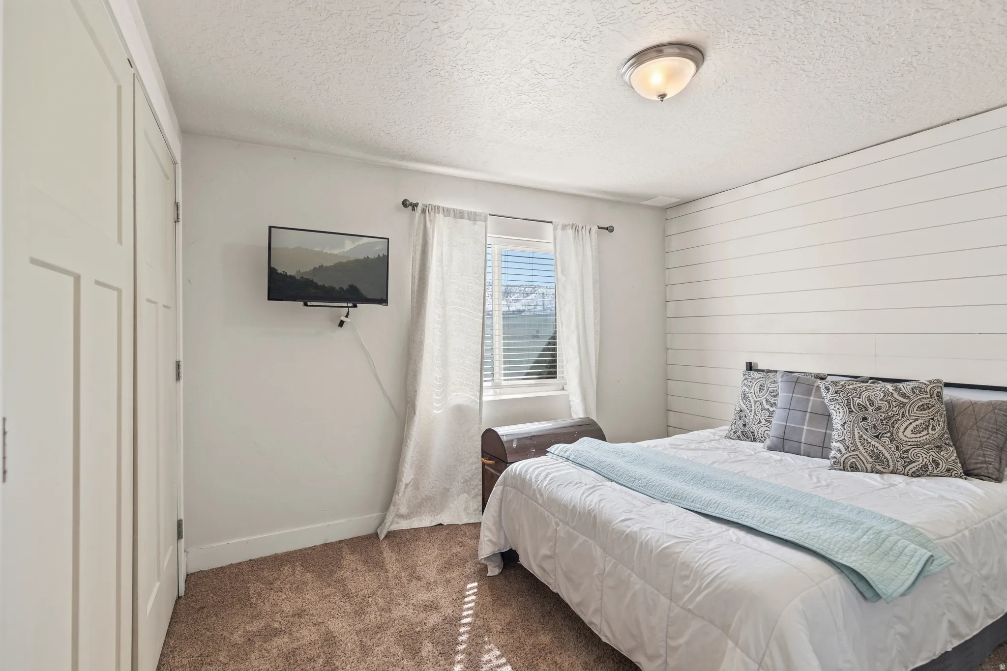 Bedroom featuring textured carpet flooring, white shiplap accent wall, light-filtering window treatments, and a semi-flush mount ceiling light fixture
