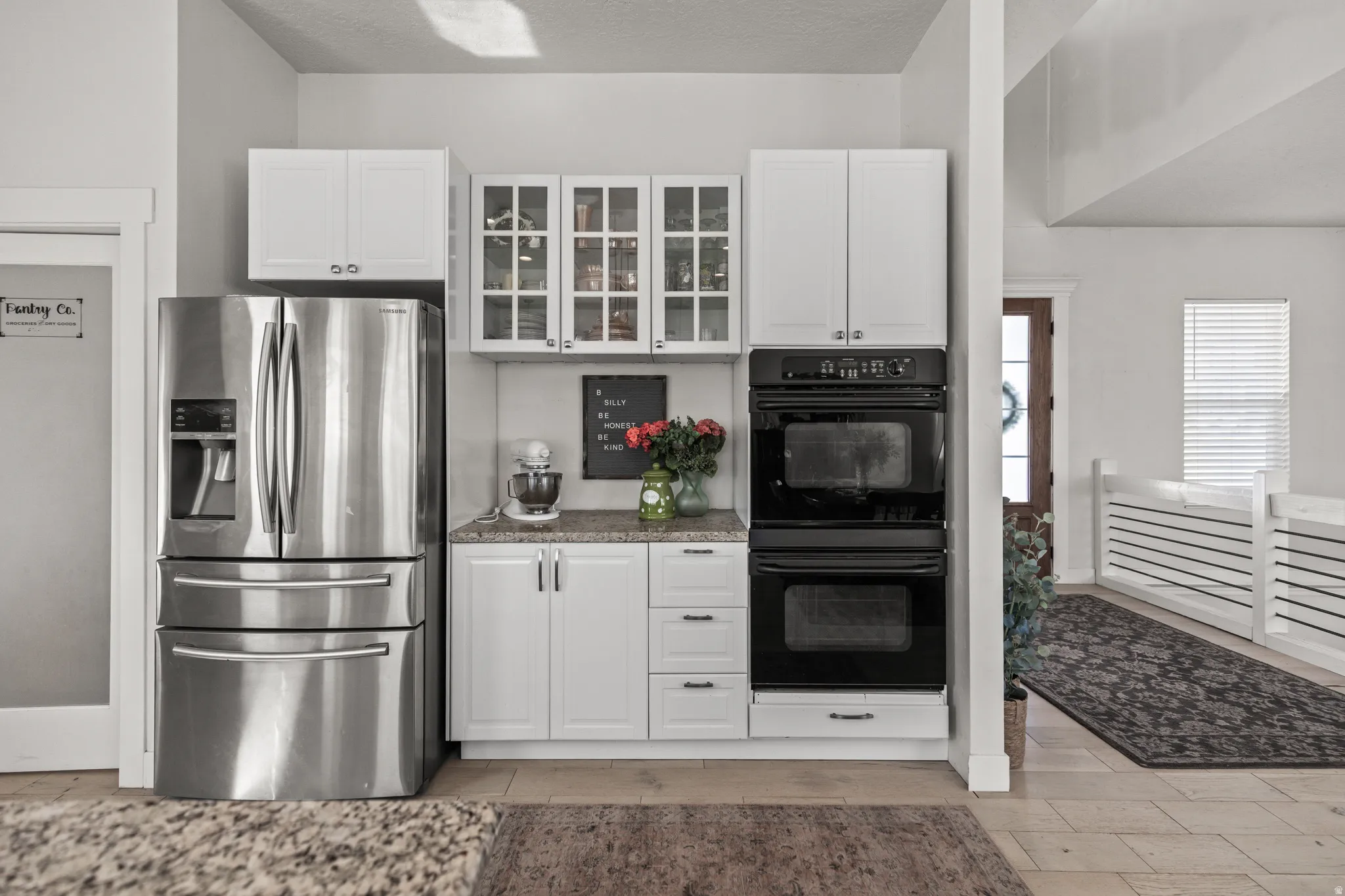 Kitchen features stainless steel refrigerator, double wall ovens, granite-style countertops, white cabinetry with glass-front uppers, and wood-finish flooring