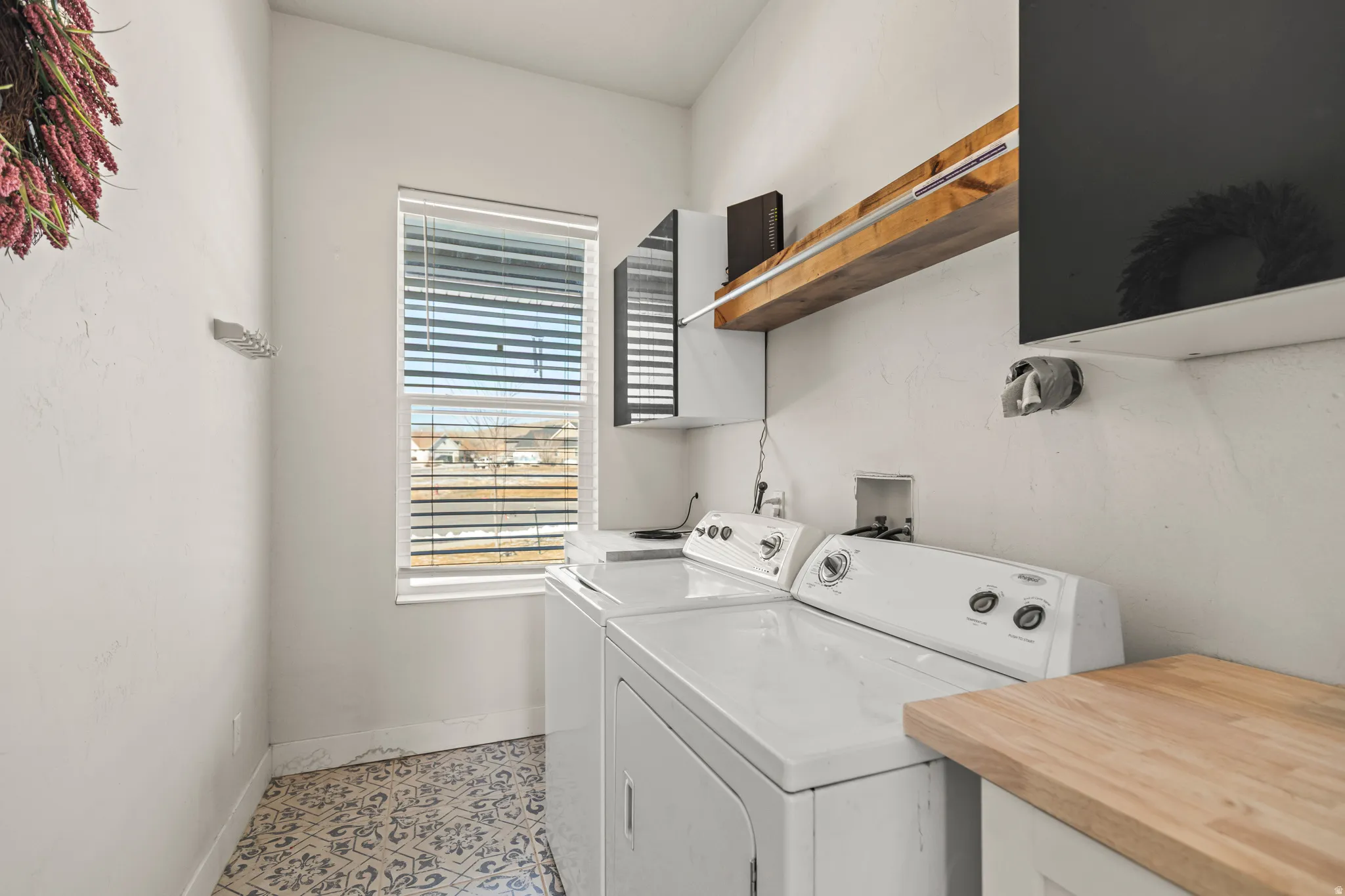 Functional laundry area featuring white walls, patterned tile flooring, a window with blinds, an overhead wooden shelf, and a light wood-finish countertop