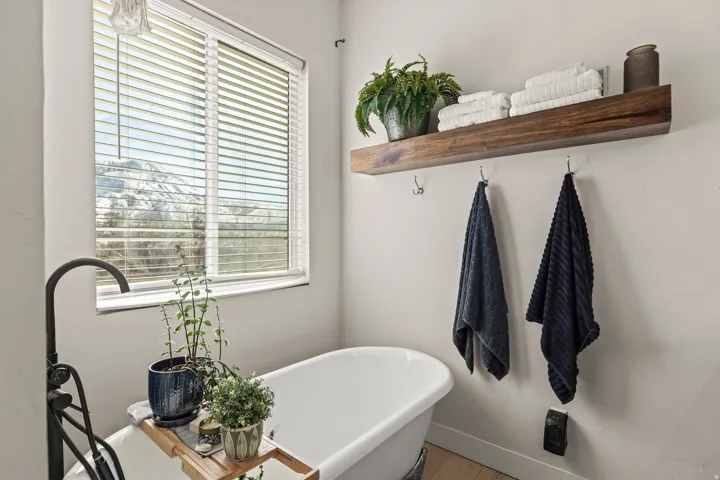 Freestanding soaking tub with a floor-mounted faucet, complemented by a rustic wood floating shelf and wall-mounted towel hooks