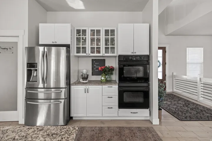 Kitchen features stainless steel refrigerator, double wall ovens, granite-style countertops, white cabinetry with glass-front uppers, and wood-finish flooring